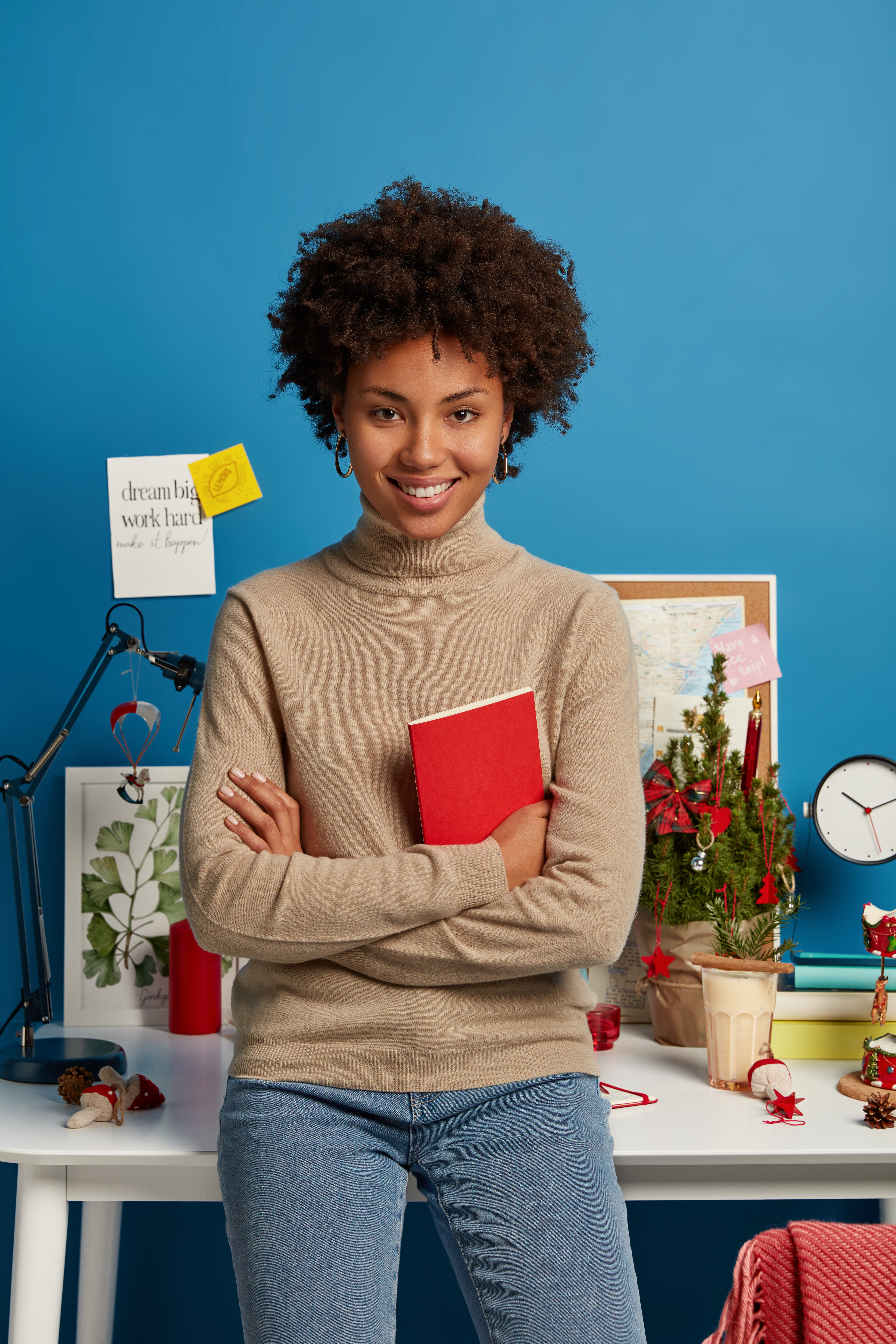 Vertical shot of pleased curly haired female teacher prepares for lessons at home, holds red textbook, poses against desktop, wears turtleneck and jeans, isolated on blue wall. Student in studyroom